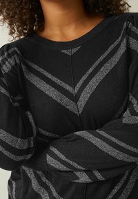 Pull en maille noire avec des rayures horizontales argentées et une finition texturée. Il présente un col rond et des manches bouffantes.