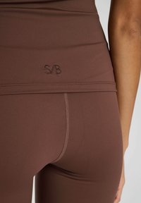 Brązowe legginsy sportowe z wysokim stanem, przeszyciami oraz wytłoczonym logo "S/B" na plecach. Gładki, elastyczny materiał.