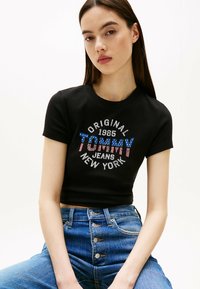 Czarny, prążkowany, krótki top crop z logo w wielu kolorach "TOMMY JEANS ORIGINAL 1985 NEW YORK" z czerwonymi i niebieskimi akcentami.