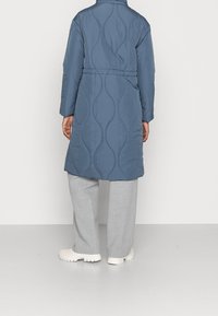 Abrigo largo acolchado azul con cintura ajustada, que presenta un cuello alto y mangas largas, usado sobre pantalones grises y calzado blanco.