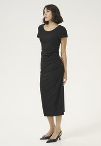 Robe noire ajustée avec des manches courtes, un col rond et un détail froncé sur le côté. Associée à des talons aiguilles noirs avec des boucles.