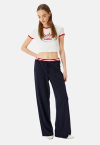 Hvid cropped t-shirt med røde kanter og grafisk tekst; marineblå bukser med vide ben og stribet talje. Glat stof, afslappet pasform.