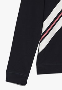 Manga de sudadera de color marino con una franja diagonal blanca y una franja roja más delgada que atraviesa la franja blanca.