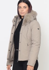 Giacca imbottita beige con cappuccio in pelliccia sintetica, chiusura con zip e bottoni a pressione, tasche laterali e un maglione nero a trama sotto.