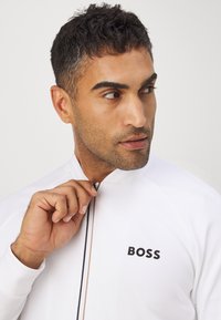 Witte zip-up jas met een hoge kraag, voorzien van een subtiele textuur en een zwart logo "BOSS" op de borst. Model houdt de ritsring vast.