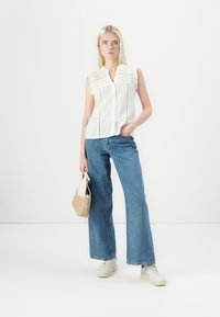 Weiße ärmellose Bluse mit strukturiertem Stoff und Knopfverschluss, kombiniert mit hellblauen weit geschnittenen Jeans und weißen Sneakers. Hält eine geflochtene Tasche.