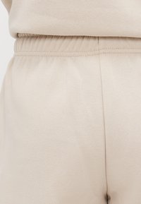 Close-up van een beige elastische tailleband op zachte stoffen broek met zichtbare naad en subtiele textuur.