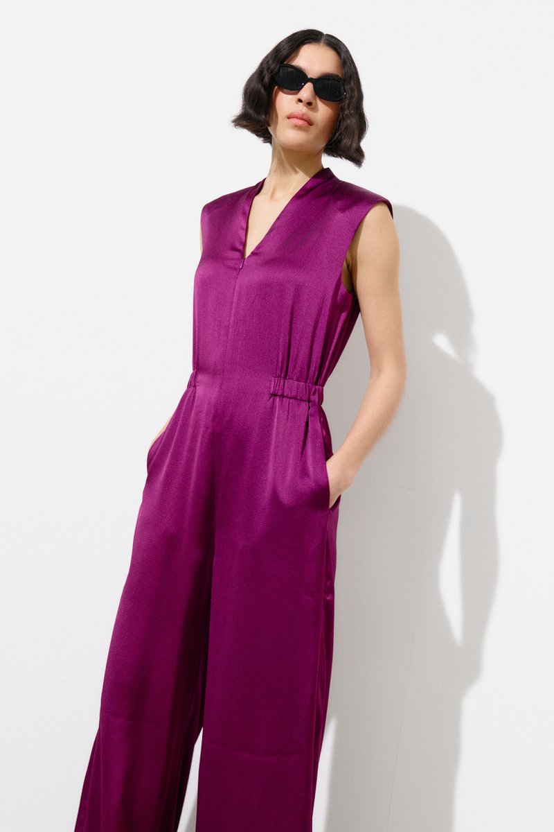 Femme aux cheveux courts foncés portant des lunettes de soleil noires et une combinaison violette sans manches à jambes larges, mains dans les poches, debout sur fond blanc.