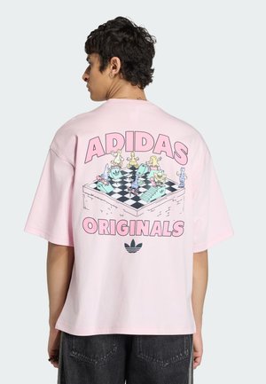Osoba ubrana w różowy T-shirt Adidas Originals z kolorowymi figurami szachowymi na rolkach wokół unoszącej się grafiki planszy do szachów.