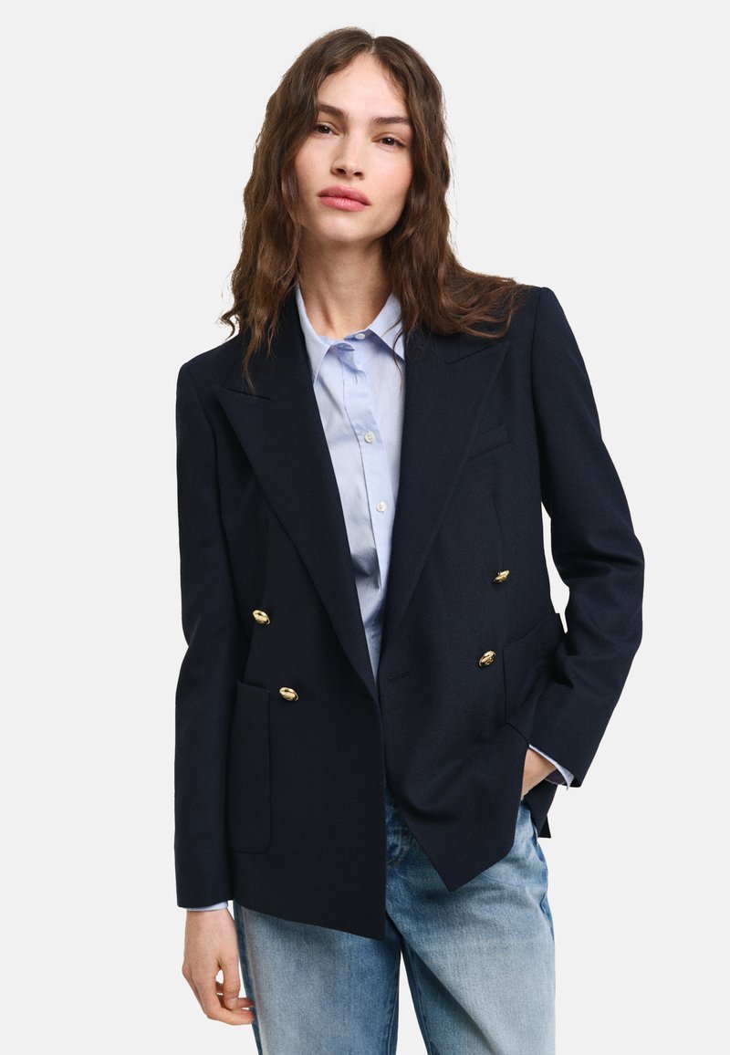 Marineblauwe dubbelgebroken blazer met gouden knopen, slim fit, met uitgesneden revers en zijzakken, gecombineerd met een lichtblauwe overhemd met kraag.