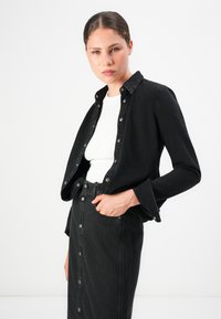 Ung kvinne iført svart denimjakke med knapper og skjørt over en hvit topp, poserer med en hånd i skjørtlommen mot hvit bakgrunn.