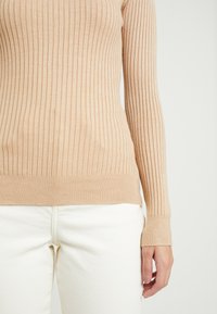 Beige žebrovaný svetr s dlouhým rukávem, nošený s kalhotami vysokého pasu v off-white barvě, ukazující torzo a pravou ruku stojící osoby.