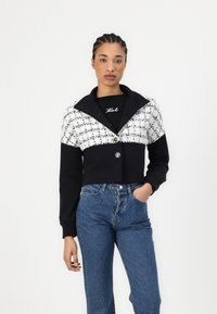 Cropped jacket featuring a black body with a white textured upper section, wide collar, and button closure, paired with high-waisted jeans.