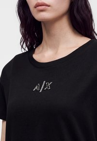 Γυναίκα με σκούρα μαλλιά φοράει μαύρο T-shirt με μικρά γράμματα A/X από στρας στο στήθος, μπροστά σε απλό φόντο.
