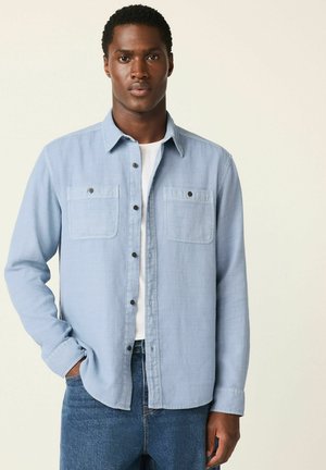 Light blue button-up shirt made of soft fabric, featuring two chest pockets, black buttons, and a collar, paired with dark denim jeans.