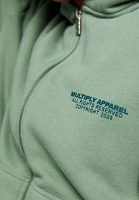 Sweat à capuche zippé vert clair en tissu doux, avec un logo bleu portant le texte "MULTIPLY APPAREL" et un avis de copyright à l'avant.