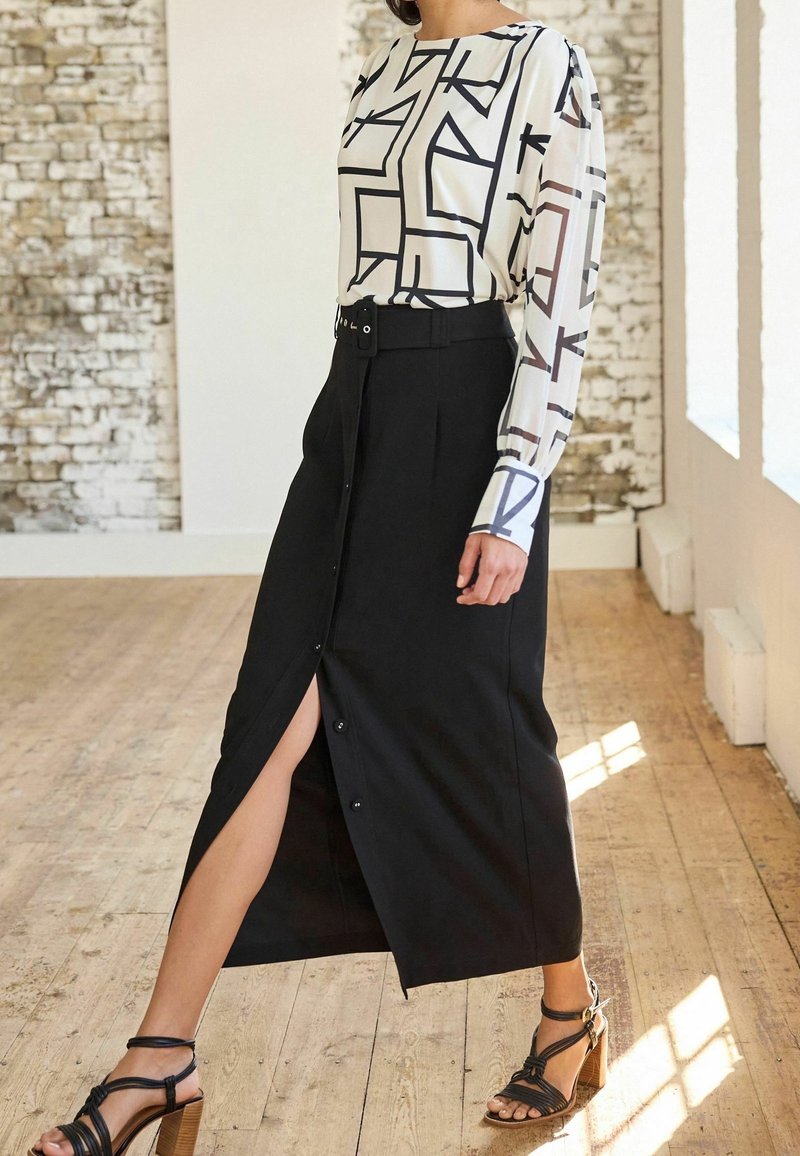 Black button-down skirt with a side slit, paired with a white top featuring a geometric black pattern. Model wears black strappy heels.