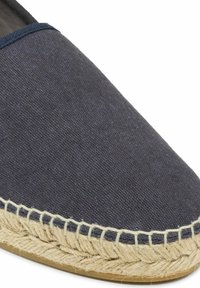 Close-up van een marineblauwe espadrille met een natuurlijke jute zool en beige stiksel langs de rand, tegen een witte achtergrond.