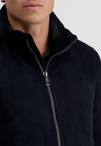 Marineblauwe, zachte suède jas met een hoge kraag en volledige ritssluiting. Textuuraccenten bij de kraag voegen detail toe aan het ontwerp.