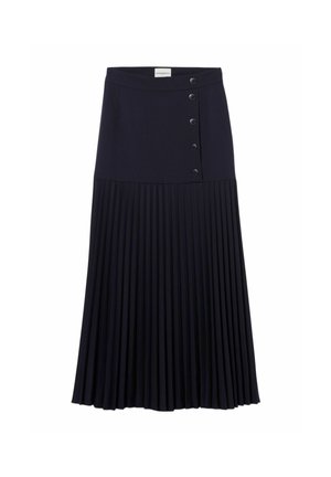 Donna che indossa un berretto beige, top nero senza maniche, gonna lunga plissettata blu scuro con bottoni e sandali dorati con cinturini, in piedi davanti a uno sfondo semplice.