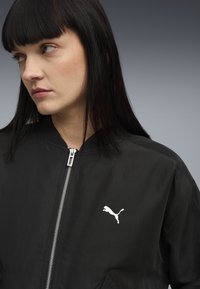 Veste bomber noire avec une fermeture éclair, un col côtelé et un logo Puma blanc sur la poitrine. Texture lisse, matériau léger.
