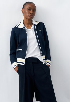 Frau mit geflochtenem Haar, die eine marineblaue Jacke mit Reißverschluss mit weißen und gelben Streifen, ein weißes Poloshirt und lockere schwarze Shorts trägt, steht mit den Händen in den Taschen.