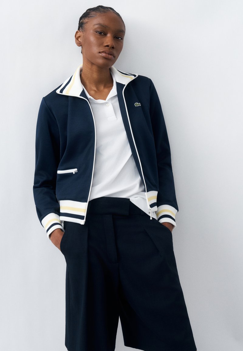 Γυναίκα με κοτσίδες, που φοράει μπλε ζακέτα με φερμουάρ με λευκές και κίτρινες ρίγες, λευκό πόλο και φαρδιά μαύρα σορτς, στέκεται με τα χέρια στις τσέπες.