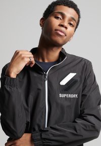 Chaqueta rompevientos negra con puños elásticos, cierre de cremallera y diseño de logo bordado. Textura suave con un aspecto deportivo y ligero.