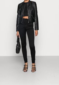 Donna che indossa una giacca di pelle nera cropped, un top nero corto, pantaloni neri a vita alta, tacchi neri con strappi, e tiene in mano una piccola borsa nera.