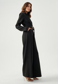 Zwarte jumpsuit met lange mouwen, v-hals en wijde pijpen. Voorzien van een riem met een ronde gouden sluiting en zijzakken. Gladde stofstructuur.