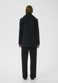 Tamsiai vilnos mišinys paltas su įpjautu apykakle, rodomas iš nugaros, derinamas su juodomis plačiomis kelnėmis ir auliniais batais. Lygus paviršius.