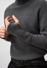 Donkergrijze geribbelde trui met een hoge kraag, met lange mouwen. Gecombineerd met een geweven zwarte riem, wat een getextureerde en gelaagde uitstraling geeft.