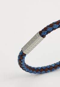 Fletet armbånd med blå tekstil og brune læderelementer, med en sølv rektangulær metalspænde mærket "GUESS."