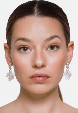 Kobieta o jasnej cerze i brązowych oczach nosi wiszące kolczyki z przezroczystymi, białymi, w kształcie liści zawieszkami.
