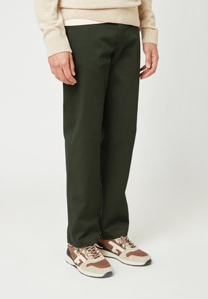 Man die donkere groene rechte broek draagt en beige, bruine en witte sneakers, staande tegen een witte achtergrond.