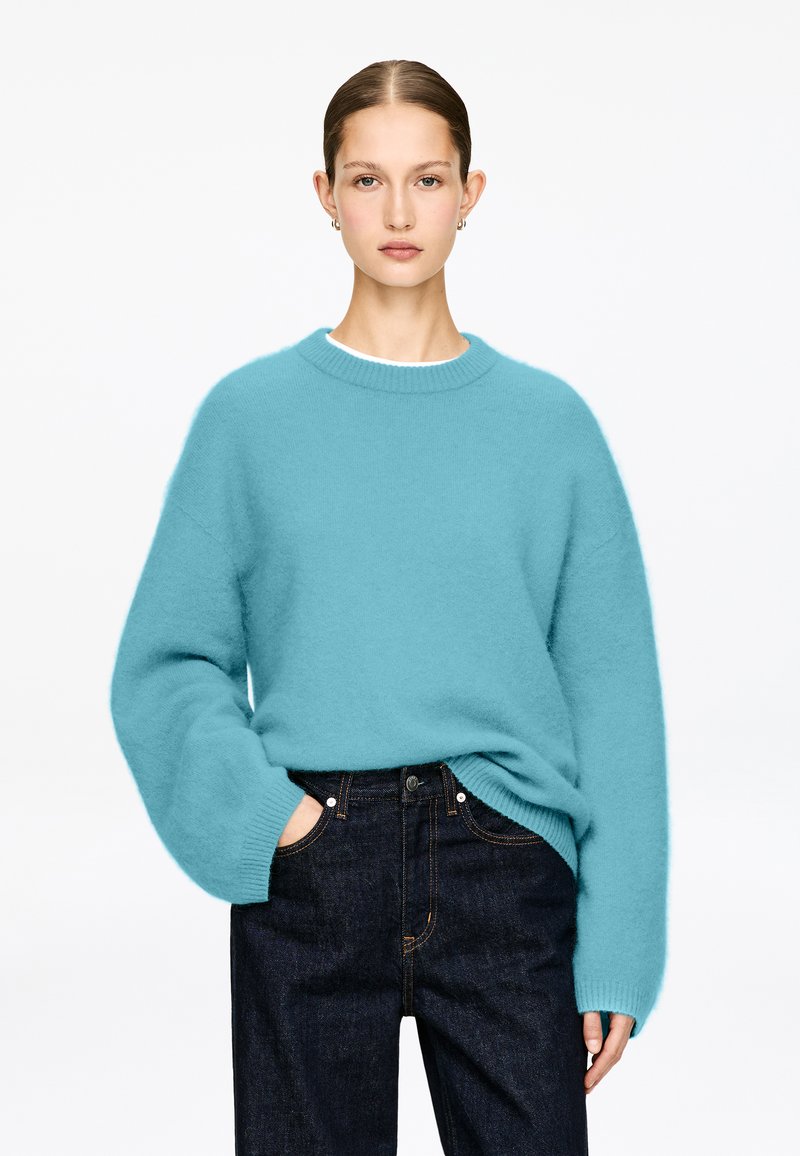 Pull surdimensionné turquoise en matière duveteuse, col rond et épaules tombantes, associé à un jean denim foncé. Design épuré.