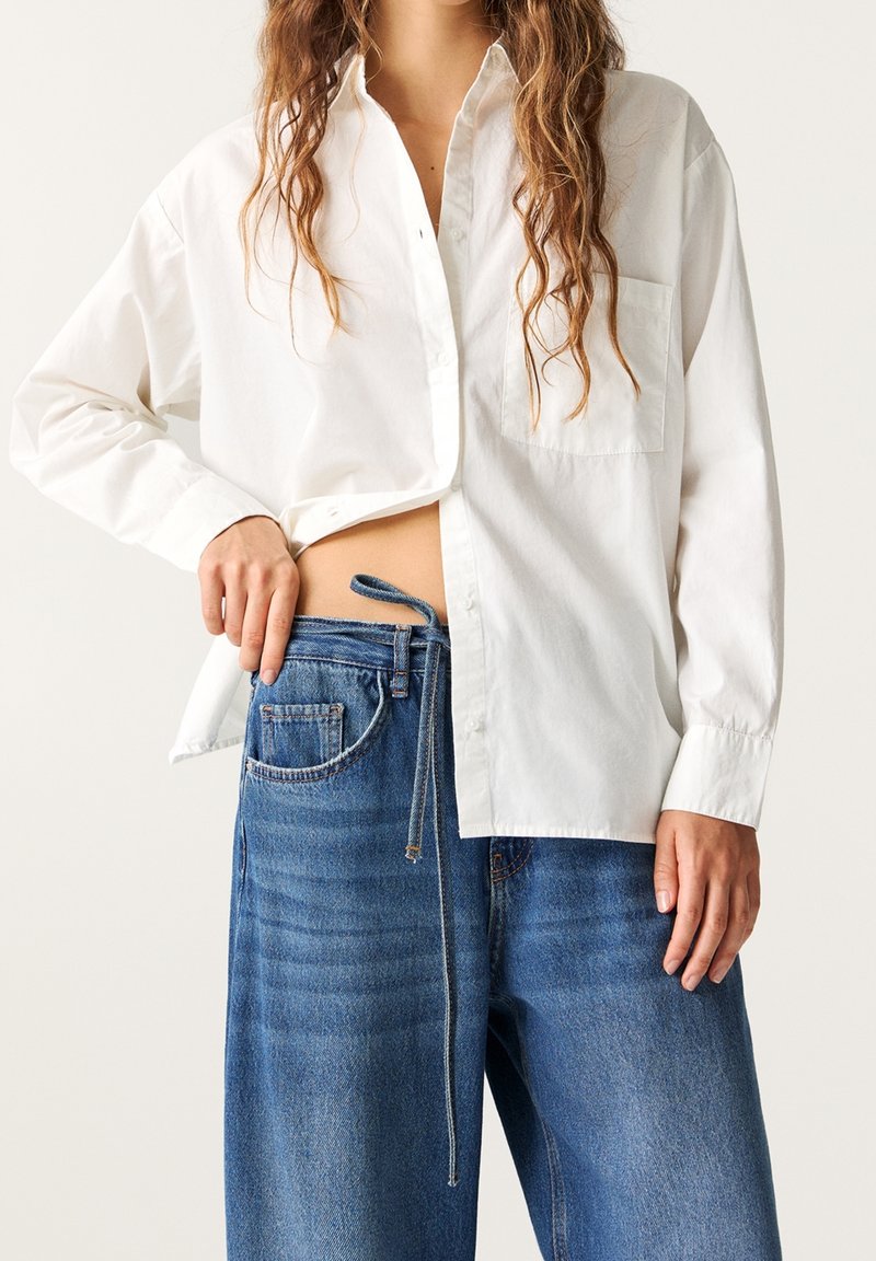 Chemise blanche à boutons avec une seule poche poitrine, associée à un jean en denim bleu coupe décontractée avec un cordon de serrage.