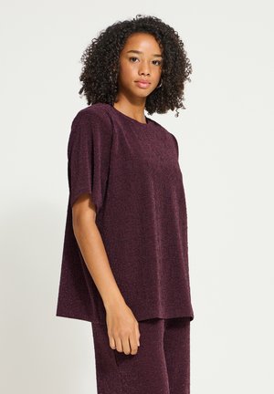 Un mannequin aux cheveux bouclés pose sur un fond uni, vêtu d’un haut ample à manches courtes couleur bordeaux et d’un pantalon assorti.