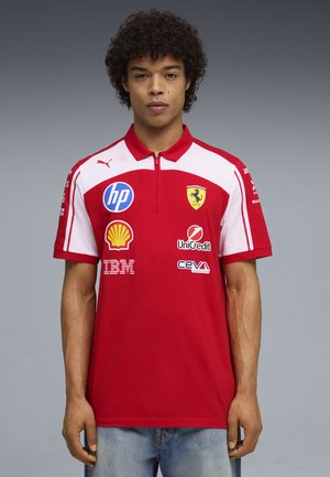Młody mężczyzna z kręconymi włosami, ubrany w czerwoną i białą koszulkę wyścigową Ferrari z logo HP, Shell, UniCredit, IBM i CEVA.