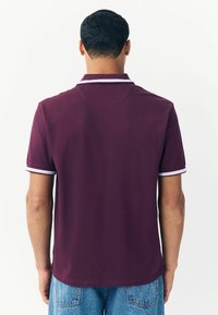 Polo di colore borgogna con colletto classico, maniche corte e accenti in bianco. Il tessuto appare liscio con una leggera texture.