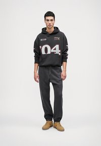 Melnas kokvilnas kapuce ar logo un grafiku '04'; savienota ar pelēkiem sporta biksēm. Modelis valkā gaiši brūnas kedas, stāvot pret tukšu fonu.