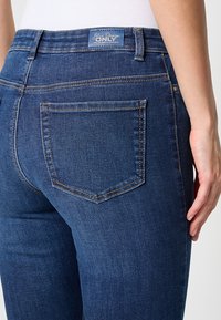 Tumed sinised denim püksid kitsas lõikega, millel on bränditud vöökoht ja üks tagatasku, millel on peened tikandite detailid.