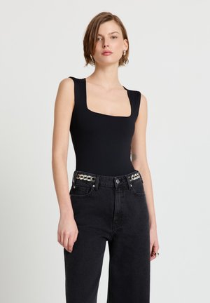 Femme aux cheveux courts portant un haut noir sans manches à encolure carrée et un jean noir taille haute avec un détail de chaîne argentée aux passants de ceinture.