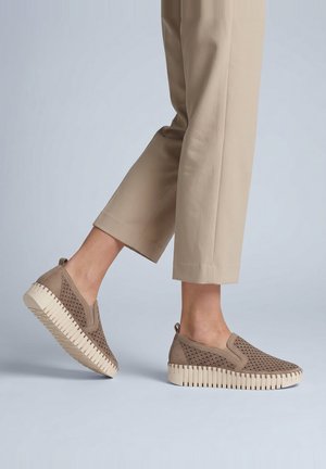 Person iført beige bukser og taupe slip-on sko med tykke hvide rillede såler på en lysegrå gulv.