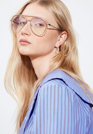 Junge Frau mit langen blonden Haaren trägt große Fliegerbrillen, silberne Creolen und ein blau gestreiftes Hemd und schaut leicht zur Seite.