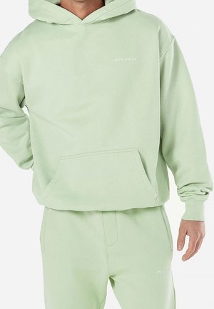 Sudadera verde claro extragrande con bolsillo frontal, puños acanalados y un logo minimalista. Hecha de un material suave, combinada con pantalones jogger a juego.