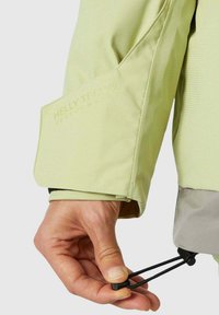 Hellgrüne Helly Tech Jacke Nahaufnahme, mit einem Bündchen mit Logo, verstellbarem Kordelzug und strukturiertem Stoff. Robustes Design für den Einsatz im Freien.