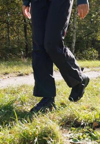 Botas de senderismo negras con suelas texturizadas y punteras reforzadas; combinadas con pantalones oscuros, caminando por un sendero natural cubierto de hierba.
