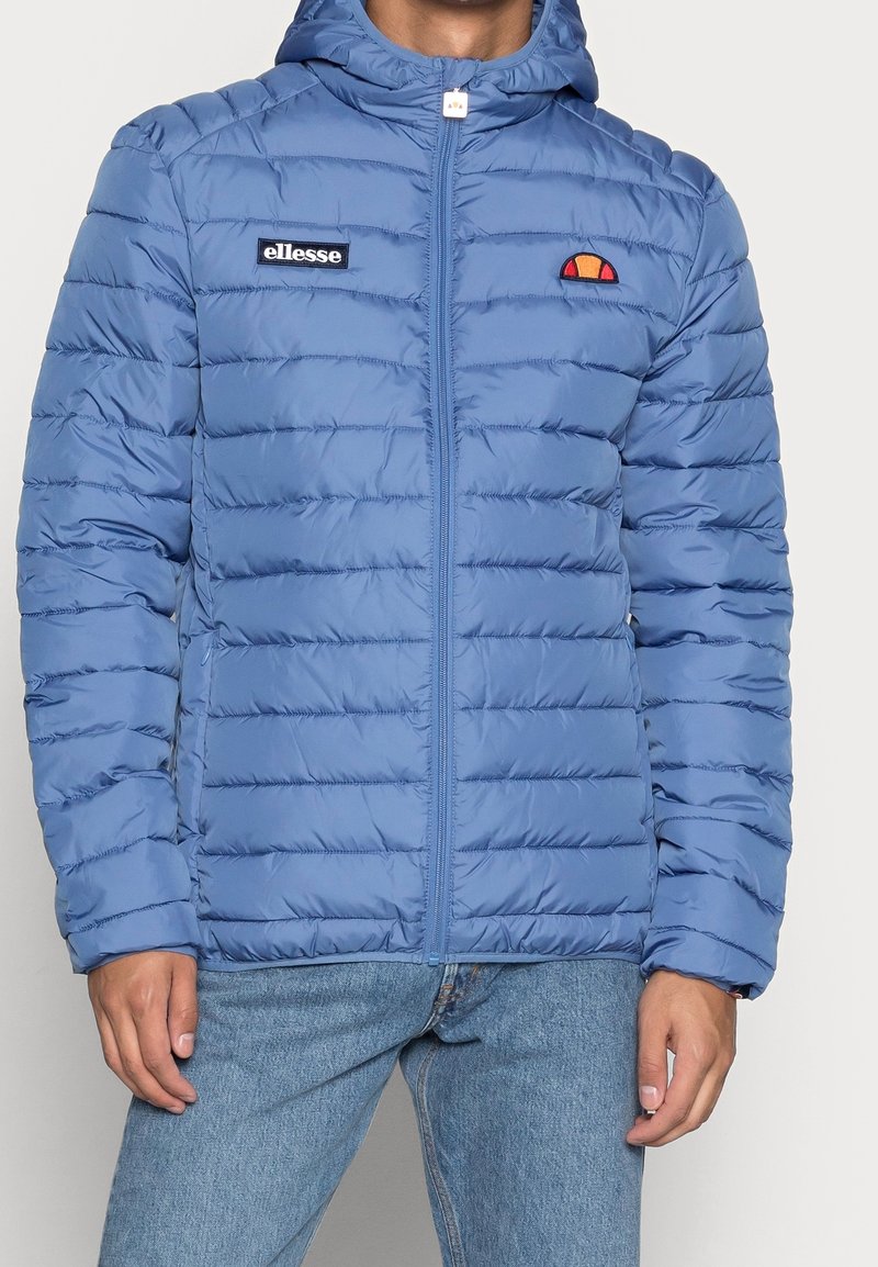 Ljusblå vadderad jacka med huva, med vertikala sömdetaljer och broderade logotyper på bröstet. Dragkedja framtill, slät textur.