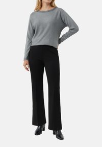 Kobieta ubrana w szary top z długim rękawem, czarne szerokie spodnie i czarne botki za kostkę, stoi z jedną ręką na biodrze.